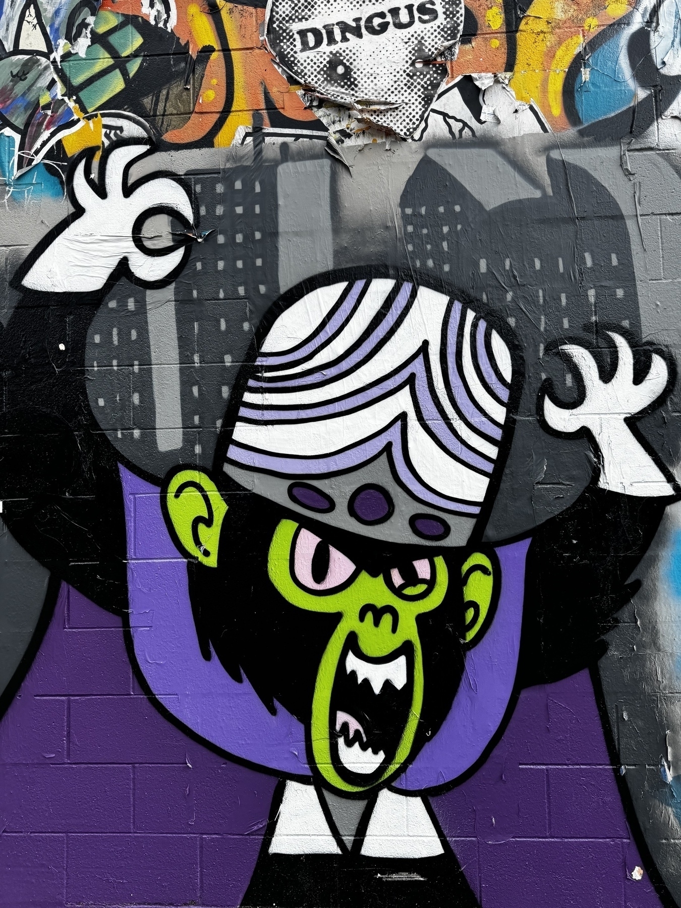 A photo of mural on a cement wall. It is focused on a depiction of the character Mojo Jojo from the Powerpuff Girls cartoon. He has his arms raised in the air and his mouth wide open. 