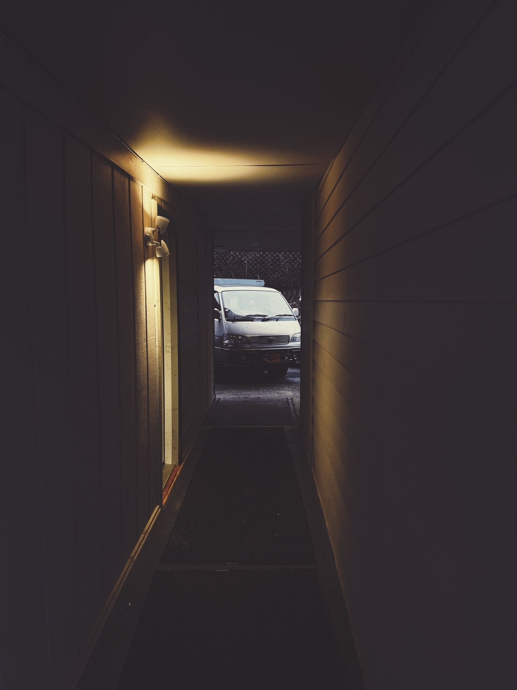 A dark photo from down hallway reveals the front of a parked Toyota HiAce van. A single light illuminates the hallway. 