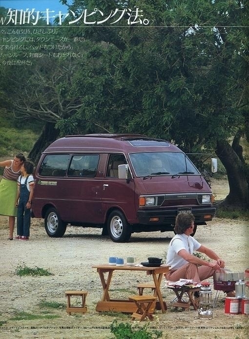 A maroon Toyota van is parked near trees. A family is setup to have a picnic. They have gear setup and a man is cooking. Nearby a woman and a child take a photo together.