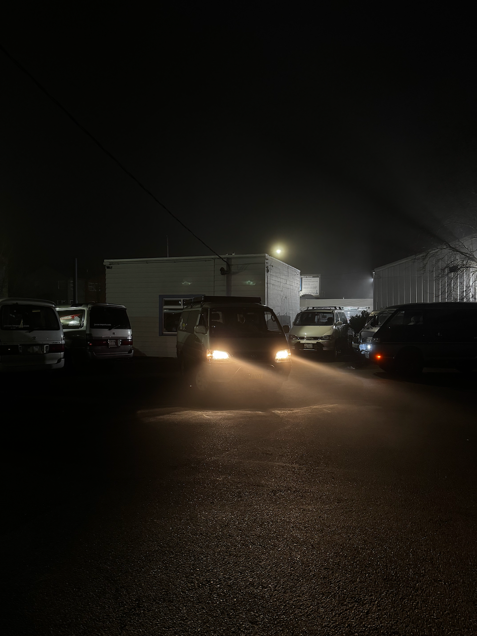 A 1998 Japanese van sits centered in the frame, parked in a lot of similar vans. Its front lights are on. It has a bull bar with fog lights mounted to it. They are off because the owner of the van got sleepy and didn't want to wait for the install to finish.