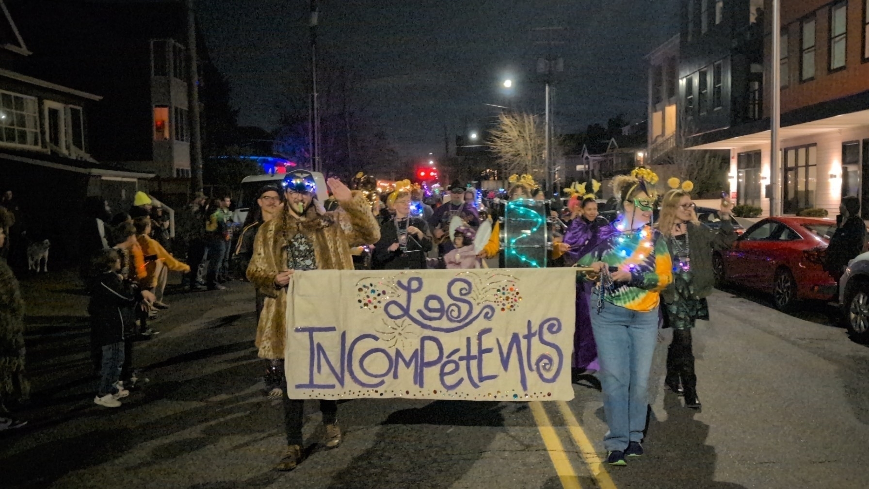 A blurry photo of a group of marchers in a Mardi Gras parade. They carry a banner between them that says Les Incompetents. They're on a dark street in the night. They're wearing yellow, green and purple some with masks and boppers on their heads. Many carry instruments.